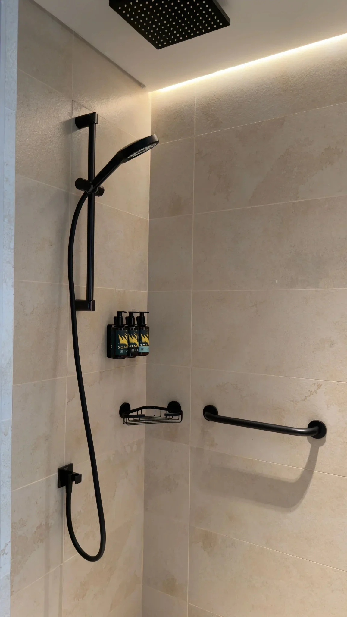 Modern hotel shower with large beige stone tiles and matte black fixtures and grab bar