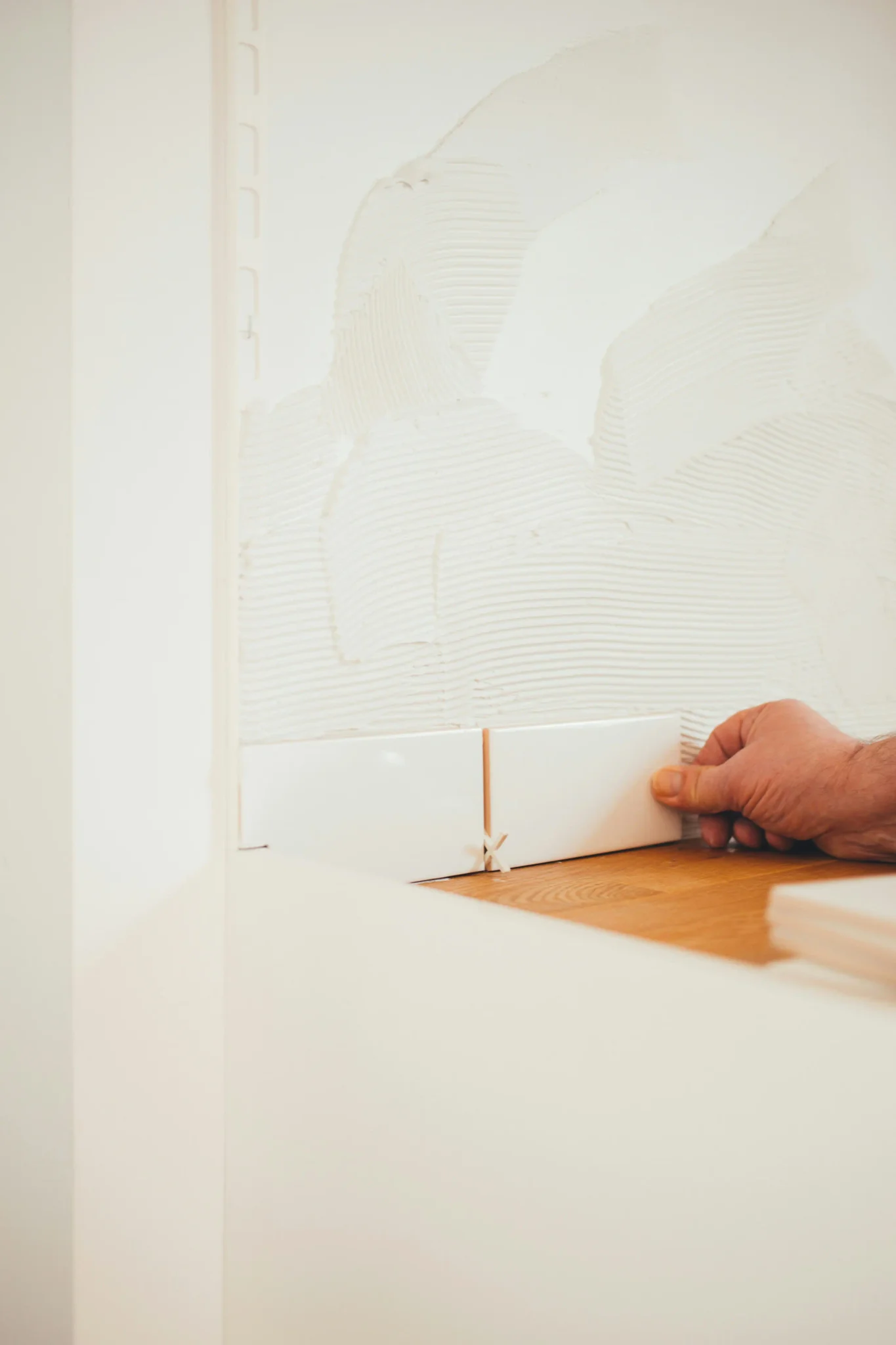 Tile installer pressing white ceramic baseboard tile onto adhesive-combed wall surface
